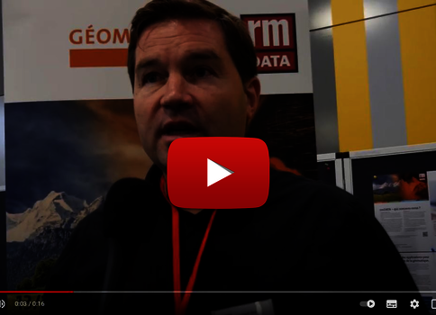 Capture d'une vidéo montrant un homme qui parle lors d'un stand d'exposition, portant un badge rouge. En arrière-plan apparaissent des affiches et logos techniques sur des panneaux multicolores.