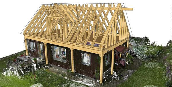 Under-construction wooden-frame house with exposed pitched roof trusses; dark exterior walls beneath the skeletal roof, set in a grassy yard with trees and an outdoor dining table and chairs to the left.