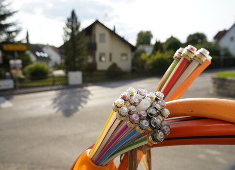 Mehrfarbige Kabelstränge mit abisolierten Metallenden liegen auf einer orangefarbenen Oberfläche; im Hintergrund sieht man ein Vorortviertel mit Häusern und Bäumen.