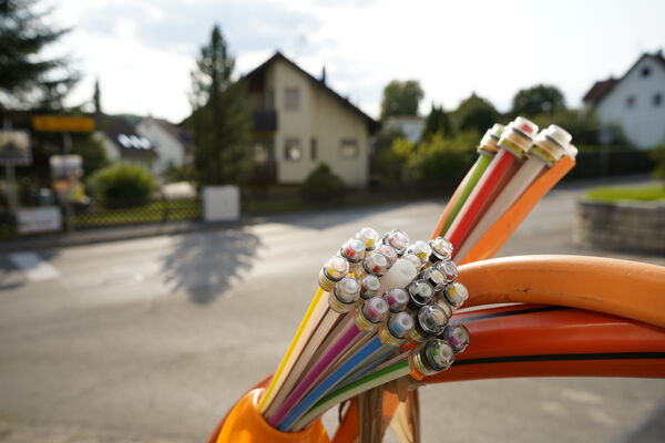Mehrfarbige Kabelstränge mit abisolierten Metallenden liegen auf einer orangefarbenen Oberfläche; im Hintergrund sieht man ein Vorortviertel mit Häusern und Bäumen.
