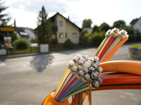 Mehrfarbige Kabelstränge mit abisolierten Metallenden liegen auf einer orangefarbenen Oberfläche; im Hintergrund sieht man ein Vorortviertel mit Häusern und Bäumen.