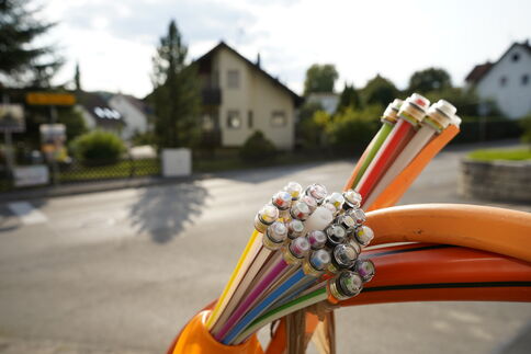 Mehrfarbige Kabelstränge mit abisolierten Metallenden liegen auf einer orangefarbenen Oberfläche; im Hintergrund sieht man ein Vorortviertel mit Häusern und Bäumen.