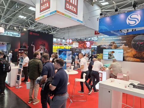 Eine Messehalle mit mehreren Messeständen, an denen Besucher an weißen Stehtischen stehen und Informationen austauschen. Große DATA-Banner hängen von der Decke, während rote Teppichflächen die Ausstellungsbereiche markieren.