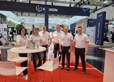 Sechs Personen in weißen Shirts stehen hinter einem runden weißen Tisch auf rotem Messeteppich. Im Hintergrund befinden sich Messestände mit Hexagon-Logo.