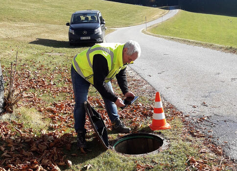 Straßenarbeiter in einer Leuchtweste kniet neben einer offenen Kanalöffnung und arbeitet am Deckel; daneben steht ein orangefarbener Warnkegel. Im Hintergrund fährt ein Auto, und Herbstlaub liegt am Straßenrand.
