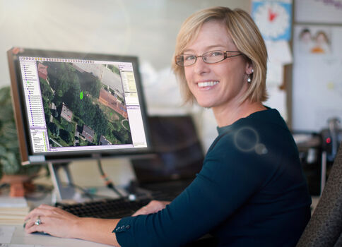 Eine Frau sitzt an einem Schreibtisch im Büro und lächelt. Auf dem Monitor ist eine Luftaufnahme einer Stadt mit Gebäuden, Straßen und Grünflächen zu sehen.