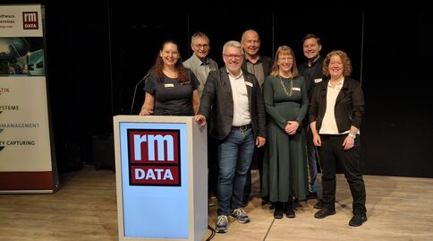 Gruppe von sieben Personen posiert hinter einem Podest mit dem rm DATA-Logo bei einer Firmenveranstaltung; alle tragen geschäftliche Kleidung und lächeln in die Kamera, während im Hintergrund Banner und eine dunkle Bühne zu sehen sind.
