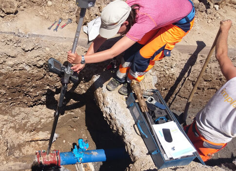 Two construction workers in a trench work on a blue water pipe with a red valve, using tools; one wears a pink shirt and orange pants, and the other holds a tool case.