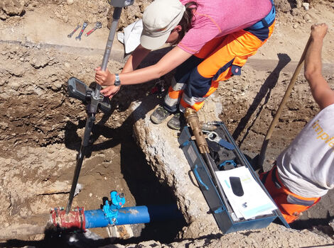 Eine Person in pinkem T‑Shirt und orangefarbener Schutzkleidung kniet in einem Graben und setzt ein Messgerät an eine blaue Rohrleitung an; daneben liegt eine tragbare Messkiste, während eine weitere Person am Rand steht.