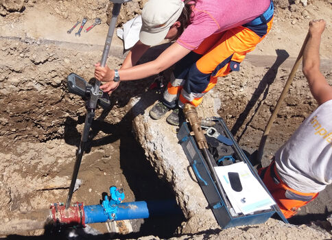 Eine Person in pinkem T‑Shirt und orangefarbener Schutzkleidung kniet in einem Graben und setzt ein Messgerät an eine blaue Rohrleitung an; daneben liegt eine tragbare Messkiste, während eine weitere Person am Rand steht.