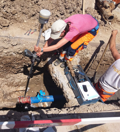 Eine Person in pinkem T‑Shirt und orangefarbener Schutzkleidung kniet in einem Graben und setzt ein Messgerät an eine blaue Rohrleitung an; daneben liegt eine tragbare Messkiste, während eine weitere Person am Rand steht.