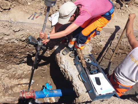 Two construction workers in a trench work on a blue water pipe with a red valve, using tools; one wears a pink shirt and orange pants, and the other holds a tool case.