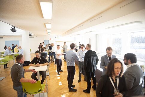 Eine Gruppe berufstätiger Menschen in einem hellen Saal beim Networking. Einige stehen im Gespräch, andere sitzen an hohen Tischen; die Szene wirkt locker und geschäftig, mit großen Fenstern an der Seite.