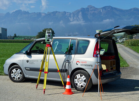 Vermessungsfahrzeug am Feldrand: Offene Heckklappe, zwei Stative mit Vermessungsgeräten, orangefarbener Verkehrskegel, Auto mit Logo "Vermessung". Hintergrund: Felder und Gebirgskette.