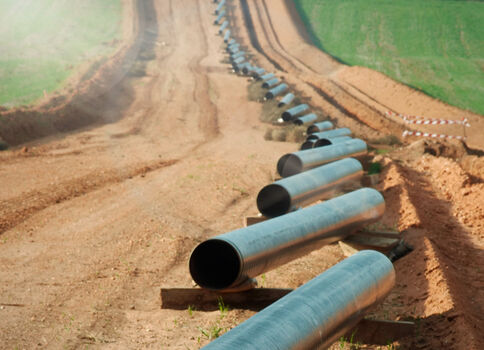 Eine lange Reihe großer Rohre liegt in einem erdigen Graben entlang eines unbefestigten Weges; Grasflächen umgeben die ländliche Baustelle, Rohre ziehen sich bis in die Ferne.