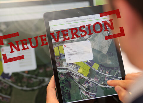 Eine Person hält ein Tablet mit Kartenansicht; auf dem Bildschirm liegt ein roter Stempel mit der Aufschrift NEUE VERSION.