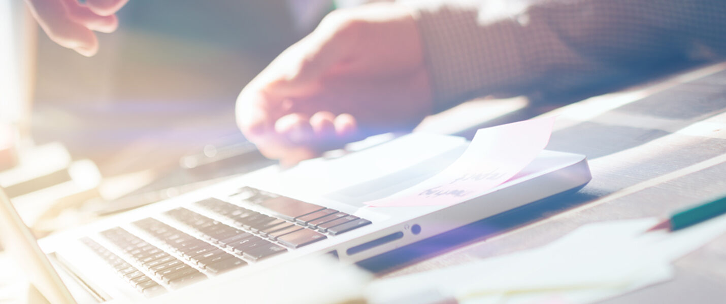 Un espace de travail moderne avec un ordinateur portable ouvert, des documents et des stylos sur le bureau, et une main posée près du clavier, éclairé par une lumière naturelle.