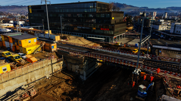 Großes Bauprojekt neben einer Eisenbahnlinie: Eine tiefe Baugrube mit Absperrungen, Bauarbeiter in orangefarbener Schutzkleidung arbeiten, Gleise verlaufen durch die Baustelle. Im Hintergrund steht ein modernes Bürogebäude mit Migros-Logo.