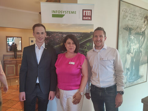 Drei Personen stehen lächelnd vor einem Banner mit den Logos ‚INFOSYSTEME‘ und ‚rm DATA‘. Links ein Mann im dunklen Anzug, Mitte eine Frau in pinkem Top, rechts ein Mann im Hemd. Es wirkt wie eine Firmenveranstaltung.