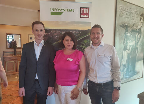 Drei Personen stehen lächelnd vor einem Banner mit den Logos ‚INFOSYSTEME‘ und ‚rm DATA‘. Links ein Mann im dunklen Anzug, Mitte eine Frau in pinkem Top, rechts ein Mann im Hemd. Es wirkt wie eine Firmenveranstaltung.