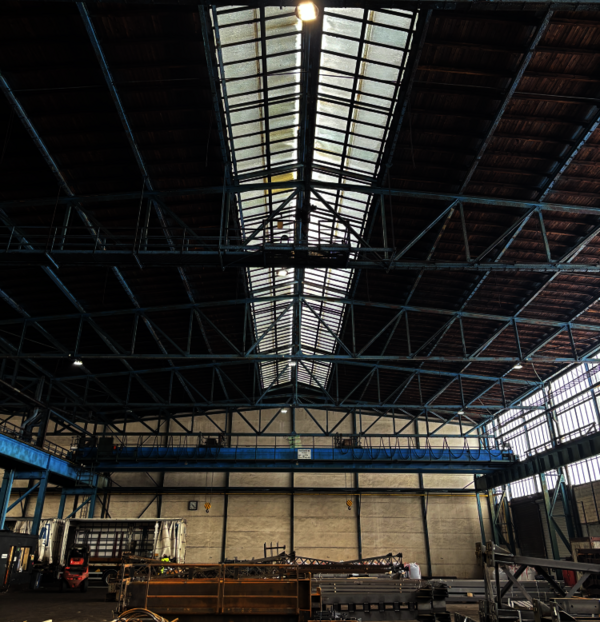 Große Lagerhalle mit hohen Decken, sichtbaren Stahlträgern und einer Dachkonstruktion aus Glasplatten; der Raum ist leer, am Rand stehen Maschinen und Paletten, hintere Wand mit Fenstern.