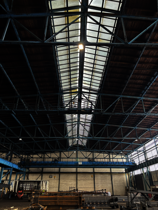 Große Lagerhalle mit hohen Decken, sichtbaren Stahlträgern und einer Dachkonstruktion aus Glasplatten; der Raum ist leer, am Rand stehen Maschinen und Paletten, hintere Wand mit Fenstern.