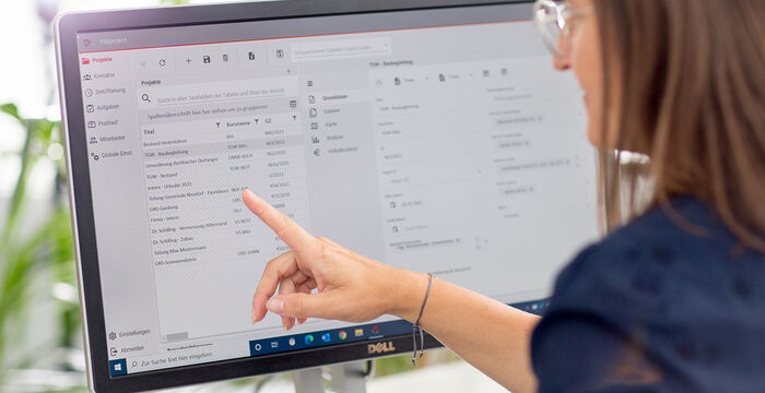 Eine Frau mit Brille sitzt vor einem Computermonitor und zeigt mit dem Finger auf den Bildschirm, der eine E-Mail-Anwendung geöffnet hat: Links befinden sich Ordner, rechts eine Liste von Nachrichten.