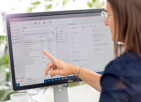A woman wearing glasses sits at a desk and points at a computer monitor displaying an open email/inbox interface, with a coffee mug nearby.