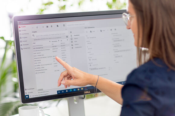 Eine Frau mit Brille sitzt vor einem Computermonitor und zeigt mit dem Finger auf den Bildschirm, der eine E-Mail-Anwendung geöffnet hat: Links befinden sich Ordner, rechts eine Liste von Nachrichten.