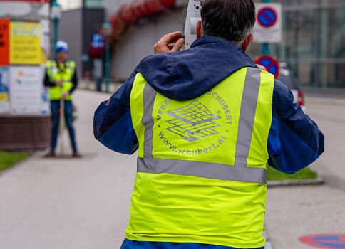 Straßenarbeiter in Leuchtwarnweste vermisst die Straße mit einem Vermessungsgerät; im Hintergrund arbeiten weitere Arbeiter neben Verkehrszeichen und einer Straßensperrung.