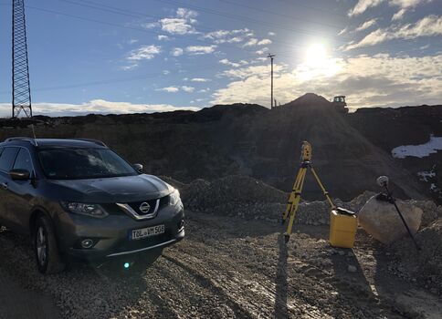 Ein grauer SUV steht auf einer unbefestigten Baupiste neben einer Vermessungsstation mit Stativ und gelber Messbox. Dahinter liegen Erdaushubhügel, Kabelmasten und die Sonne am Himmel.