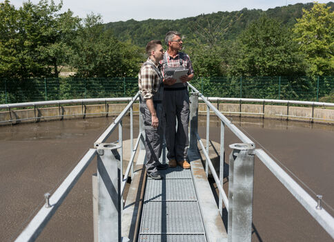Zwei Männer stehen auf einer Metallbrücke über einem runden Becken, vermutlich in einer Anlage, und schauen gemeinsam auf ein Tablet. Im Hintergrund grüne Bäume, Hecken und Hügel unter hellem Himmel.