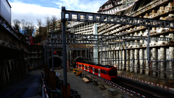 Ein roter Nahverkehrszug steht auf Gleisen in einer Baustellenszene mit Stahlrahmen, Holzbalken und Baugeräten. Die Umgebung wirkt bergbauartig; im Hintergrund sind Gebäude zu sehen, der Himmel ist blau mit Wolken.