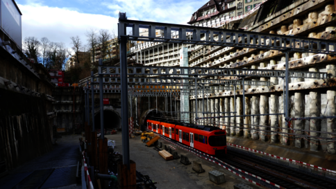 Ein roter Nahverkehrszug steht auf Gleisen in einer Baustellenszene mit Stahlrahmen, Holzbalken und Baugeräten. Die Umgebung wirkt bergbauartig; im Hintergrund sind Gebäude zu sehen, der Himmel ist blau mit Wolken.