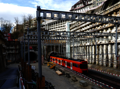 Ein roter Nahverkehrszug steht auf Gleisen in einer Baustellenszene mit Stahlrahmen, Holzbalken und Baugeräten. Die Umgebung wirkt bergbauartig; im Hintergrund sind Gebäude zu sehen, der Himmel ist blau mit Wolken.