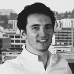 Porträt eines jungen Mannes in weißem Hemd, lächelnd zur Kamera, vor einer städtischen Skyline im Hintergrund. Schwarzweißfoto.