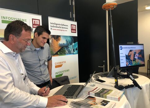 Zwei Männer stehen an einem Messestand für IT-Lösungen. Auf dem Tisch liegen Laptop, Broschüren und ein Monitor mit einer Präsentation. Hinter ihnen Banner mit rm Data und Infosysteme. Die Szene wirkt wie eine Fachmesse oder Konferenz.