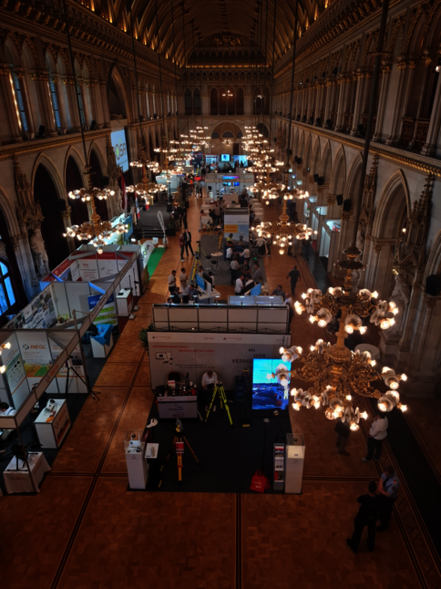 Historische Messehalle mit gewölbter Decke und prachtvollen Kronleuchtern. Zahlreiche Ausstellerstände und Besucher verteilen sich über den Saal; Menschen schauen, informieren sich und führen Gespräche.
