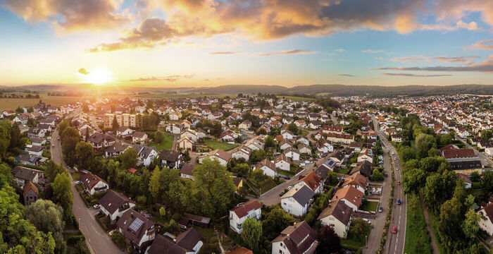 Panoramablick auf eine vorstädtische Siedlung beim Sonnenuntergang: Reihenhäuser und Straßen zwischen grünen Bäumen, Felder am Horizont, orange-gelber Himmel. Ruhiges Wohngebiet mit weitem Blick über die Stadt.