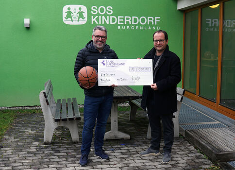 Deux hommes posent devant le bâtiment vert de SOS Kinderdorf Burgenland lors de la remise d’un grand chèque, symbolisant un don. Le chèque est présenté entre eux; des bancs et une table se trouvent à l’extérieur.
