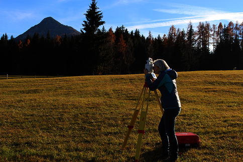 Eine Person bedient ein Theodolit auf einem Stativ zur Vermessung in einem offenen Grasfeld; im Hintergrund Wald, Berge und blauer Himmel; eine rote Transportbox liegt am Boden.