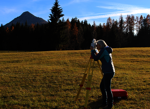 Eine Person bedient ein Theodolit auf einem Stativ zur Vermessung in einem offenen Grasfeld; im Hintergrund Wald, Berge und blauer Himmel; eine rote Transportbox liegt am Boden.