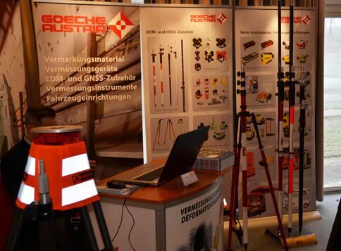 Ausstellungsstand für Vermessungstechnik: Banner mit Vermessungs- und GNSS-Zubehör, Messinstrumente und Stangen. Ein Laptop liegt offen auf dem Tisch, Hintergrund mit weiteren Messgeräten und Ausrüstung.