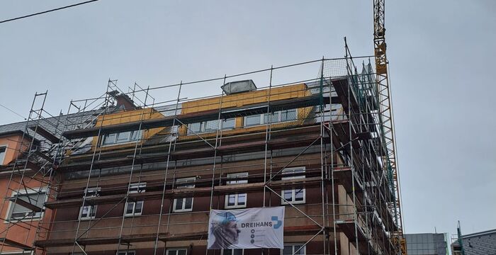 A multi-story brick building under construction, surrounded by scaffolding and a tall crane; banners with company logos hang on the scaffolding.