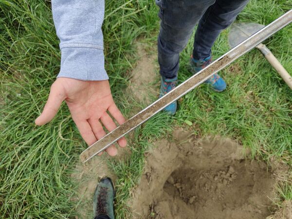 Two people stand on grass beside a dug hole, with a metal ruler laid across the rim to measure its depth.