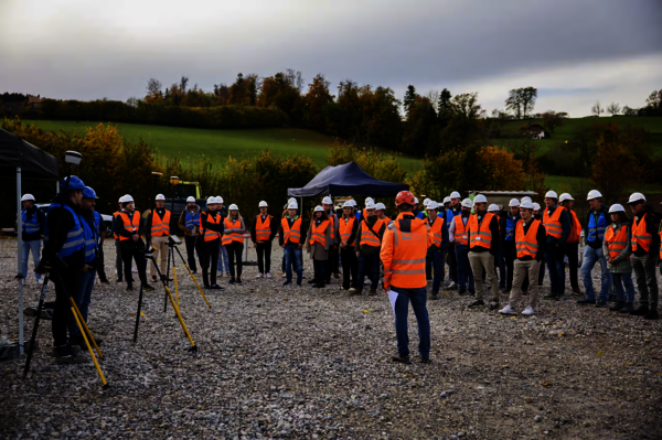 Eine Gruppe Bauarbeiter und Vermessungstechniker mit orangefarbenen Warnwesten und weißen Helmen versammelt sich in einem Kreis auf einem Kiesplatz; in der Mitte spricht eine Person, Messgeräte auf Stativen im Vordergrund, Felder und Bäume im Hintergrund.