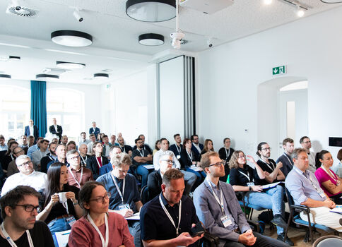 Viele Menschen sitzen in einem hellen, modernen Konferenzraum in Reihen zusammen und hören einem Vortrag zu; einige machen Notizen, tragen Namensschilder.