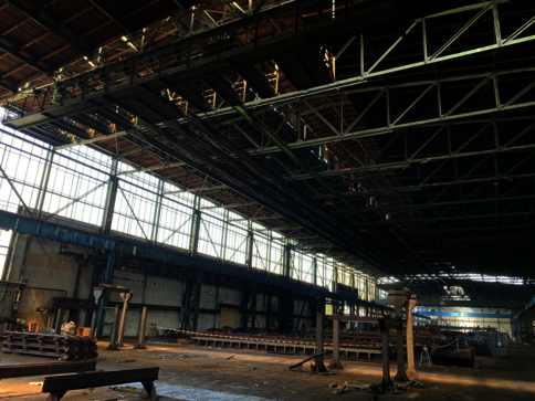 Große Industriehalle mit offener Dachkonstruktion und Stahlträgern. Links erstrecken sich Fensterreihen, durch die Tageslicht hinein fällt. Auf dem Hallenboden liegen gestapelte Metallbahnen und Bauteile; im Hintergrund sind weitere Strukturen und Gerüste sichtbar.