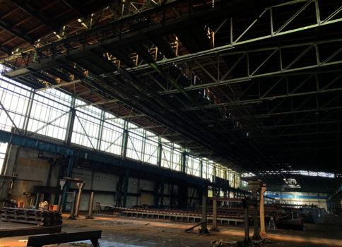 Große Industriehalle mit offener Dachkonstruktion und Stahlträgern. Links erstrecken sich Fensterreihen, durch die Tageslicht hinein fällt. Auf dem Hallenboden liegen gestapelte Metallbahnen und Bauteile; im Hintergrund sind weitere Strukturen und Gerüste sichtbar.