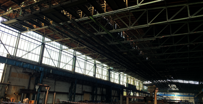 Große Industriehalle mit offener Dachkonstruktion und Stahlträgern. Links erstrecken sich Fensterreihen, durch die Tageslicht hinein fällt. Auf dem Hallenboden liegen gestapelte Metallbahnen und Bauteile; im Hintergrund sind weitere Strukturen und Gerüste sichtbar.